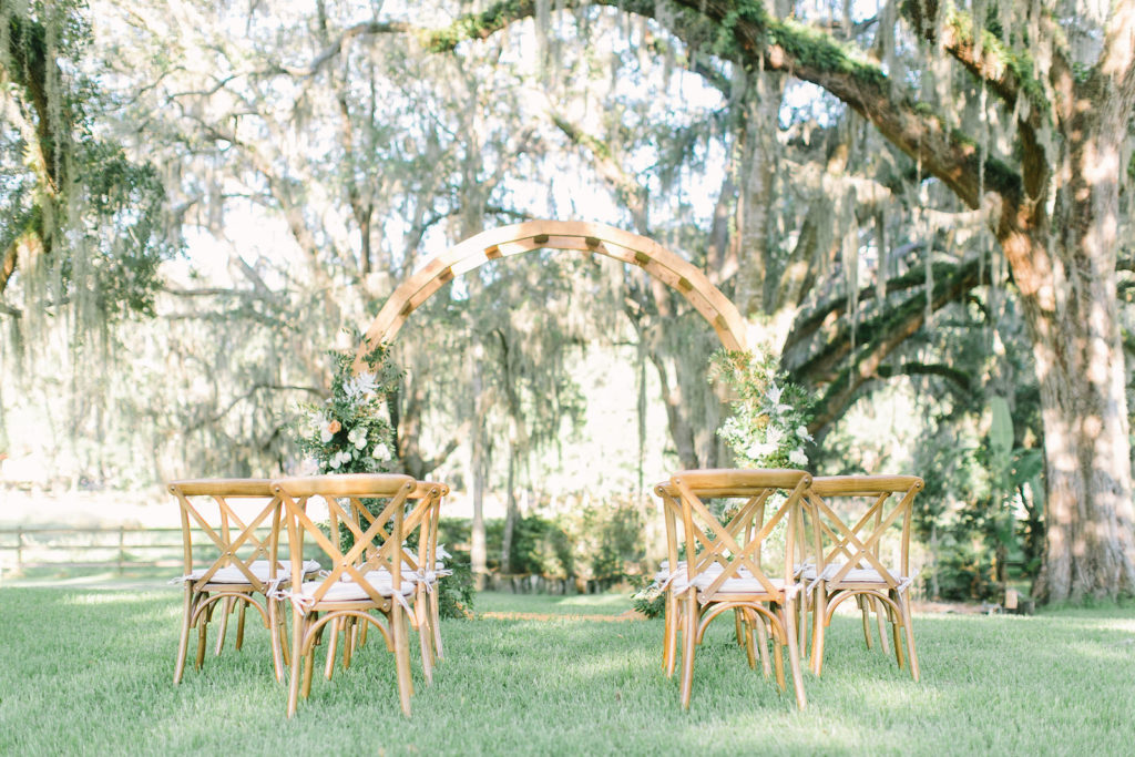 Outdoor Florida At Home Front Back Yard Wedding Ceremony | Round Wood Moon Arch Wedding Ceremony Backdrop with Natural Ivory and Peach Garland Floral Arrangements | Wood French Country Cross Back Chairs with Ivory Cushions | Intimate COVID Social Distancing Wedding