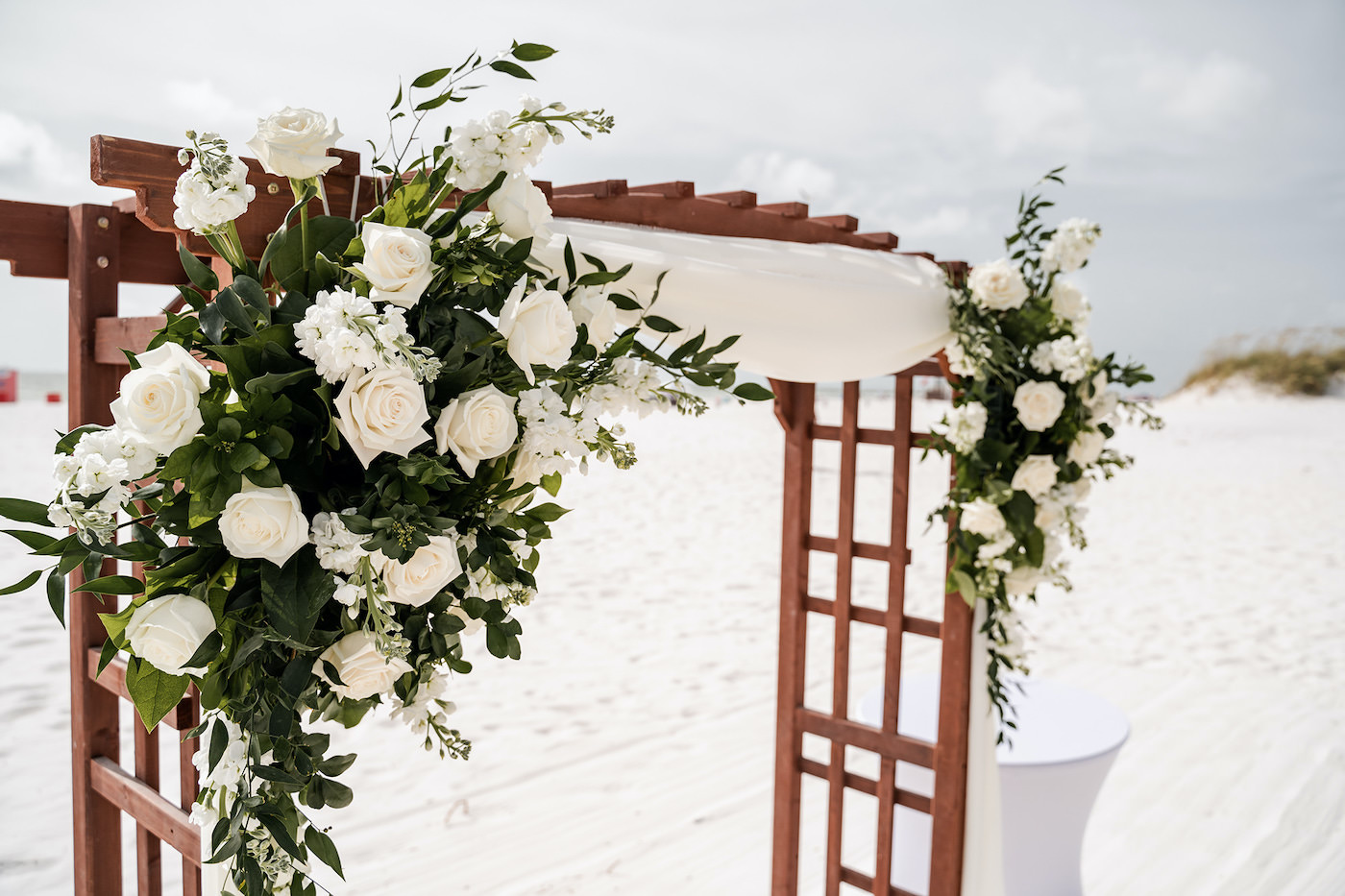 Tampa Bay Beach Wedding Ceremony, Outdoor Beachfront Ceremony on the White Powder Sand with Tropical Decor, Wooden Arch with Elegant Floral Bouquets, White Roses with Greenery and Draping | Florida Hotel and Wedding Venue Hilton Clearwater Beach