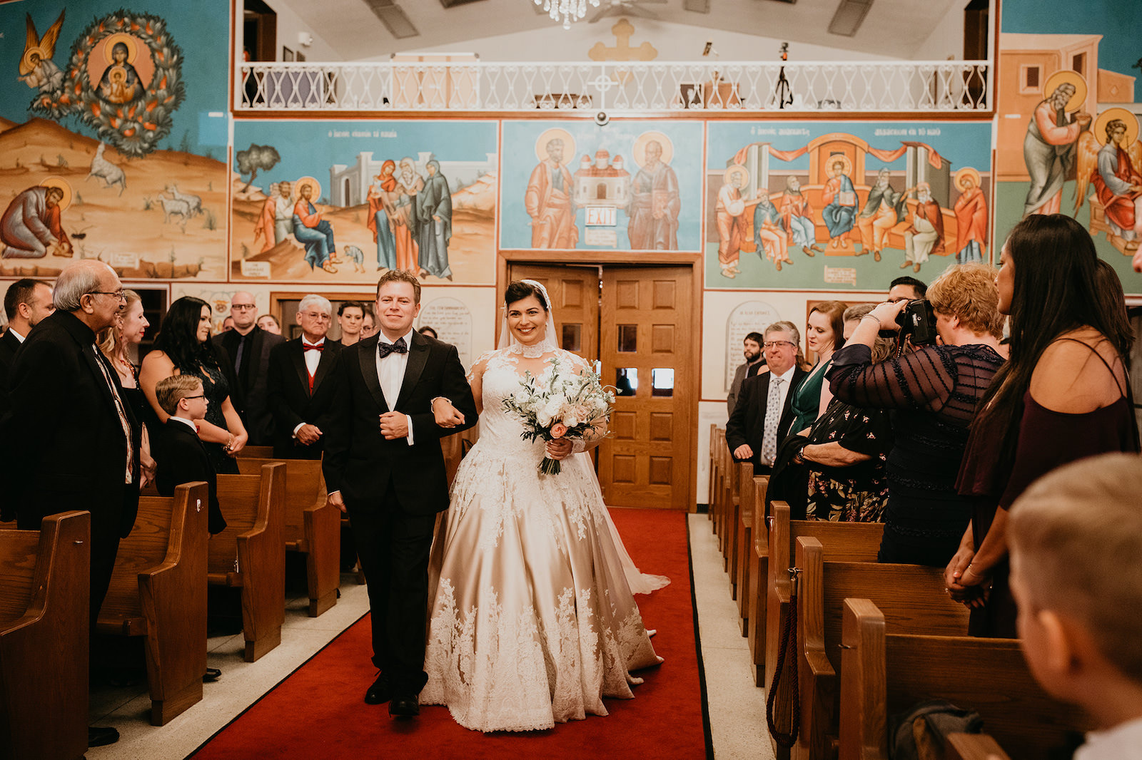St. Petersburg Florida Traditional Greek Orthodox Church Wedding Ceremony | Bride Walking Down the Aisle Wearing a Champagne and Ivory Lace Allure Bridal Ball Gown with Sweetheart Neckline and Illusion Lace Bodice and Sleeves