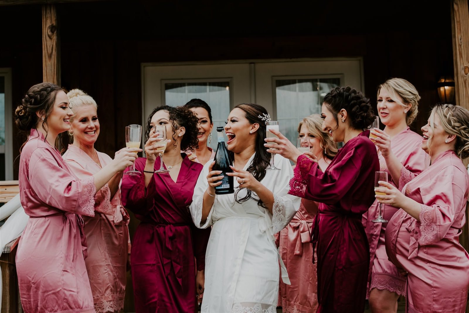 Bride and Bridesmaids Getting Ready in Pink and Burgundy Robes Popping Drinking Champagne | Femme Akoi Beauty Studio Wedding Hair and Makeup