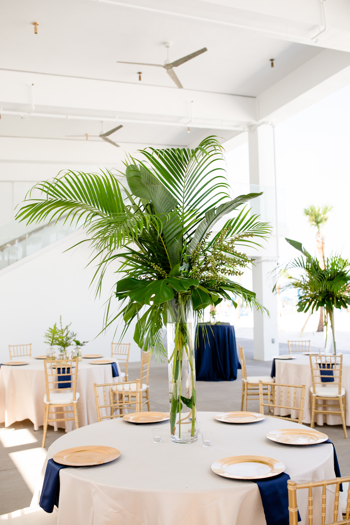 Clearwater Beach Wedding Venue Hilton Clearwater Beach | Modern Tropical Beach Outdoor Wedding Reception Terrace with White Table Linens and Navy Napkins under Gold Charger Plates | Gold Chiavari Chairs and Champagne Sash Bows and Tropical Palm Frond Leaf Floral Arrangement Centerpieces