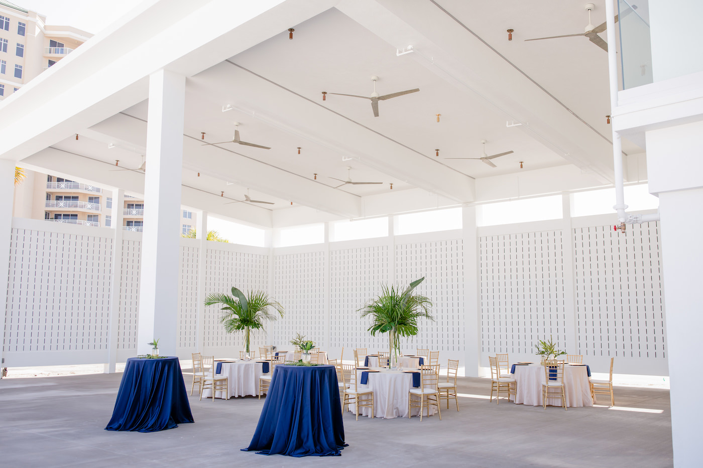 Clearwater Beach Wedding Venue Hilton Clearwater Beach | Modern Tropical Beach Outdoor Wedding Reception Terrace with White Table Linens and Navy Napkins under Gold Charger Plates | Gold Chiavari Chairs and Champagne Sash Bows and Tropical Palm Frond Leaf Floral Arrangement Centerpieces