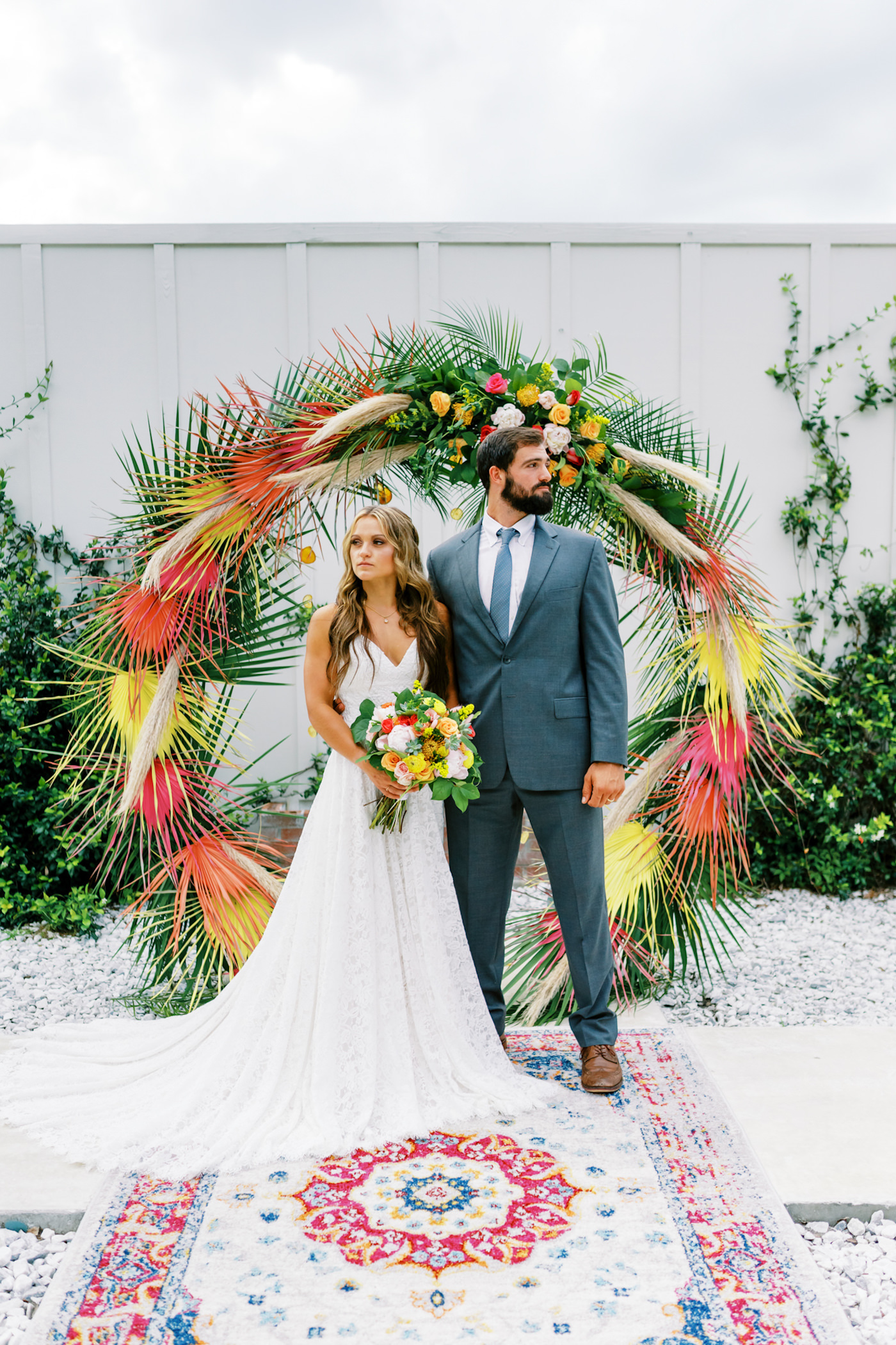 Tropical Florida Citrus Wedding Inspiration | Orange and Yellow Painted Palm Frond Leaf Round Moon Arch Ceremony Backdrop with Pampas Grass and Hanging Orange Slices | Bright Colorful Floral Arrangement with Orange and Pink Roses and Yellow Pincushion Protea | Ceremony Area Rug | Groom in Grey Suit