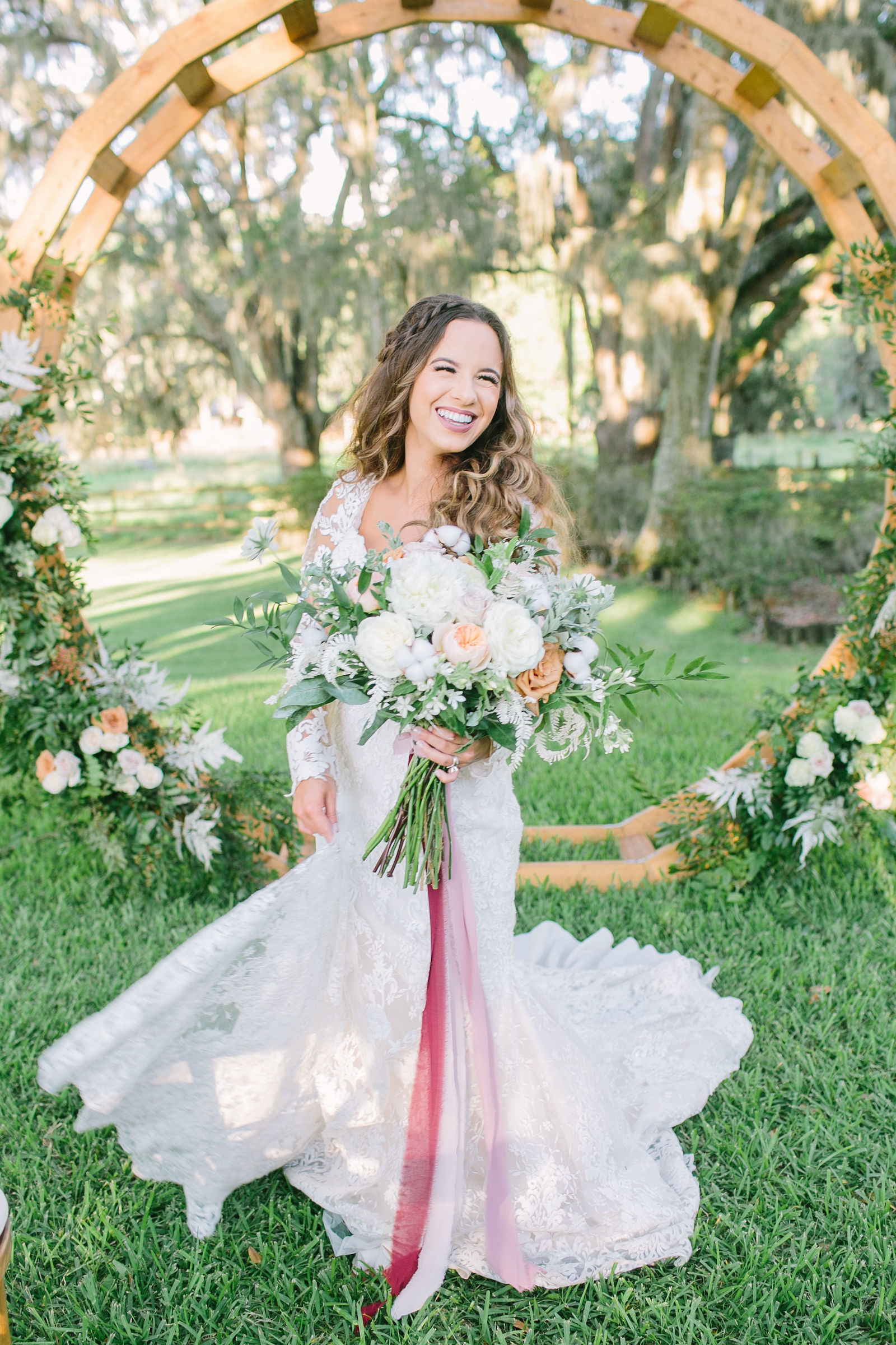 Outdoor Florida Bridal Portrait | Lace Mermaid Long Sleeve Low Back Illusion Lace Bridal Gown Wedding Dress | Ivory Champagne and Peach Natural Bouquet with Roses Astilbe and Greenery tied with Blush Pink and Mauve Ribbons | Round Wood Moon Arch Wedding Ceremony Backdrop with Natural Ivory and Peach Garland Floral Arrangements