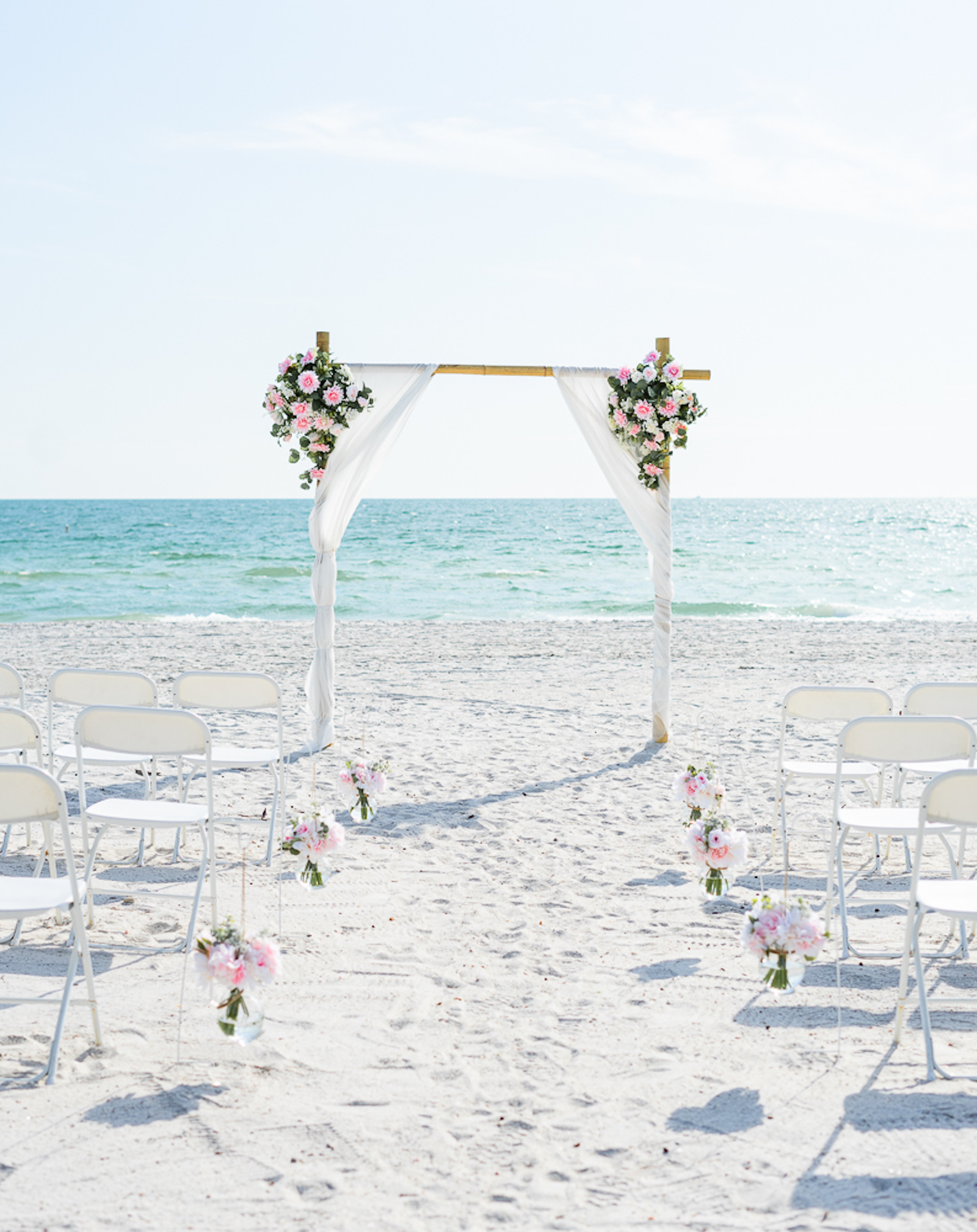 St. Petersburg Florida Beach Wedding Ceremony | Bamboo Arch Backdrop with Sheer White Draping and Blush Pink Floral Arrangements | White Ceremony Chairs with Blush Pink Aisle Flowers