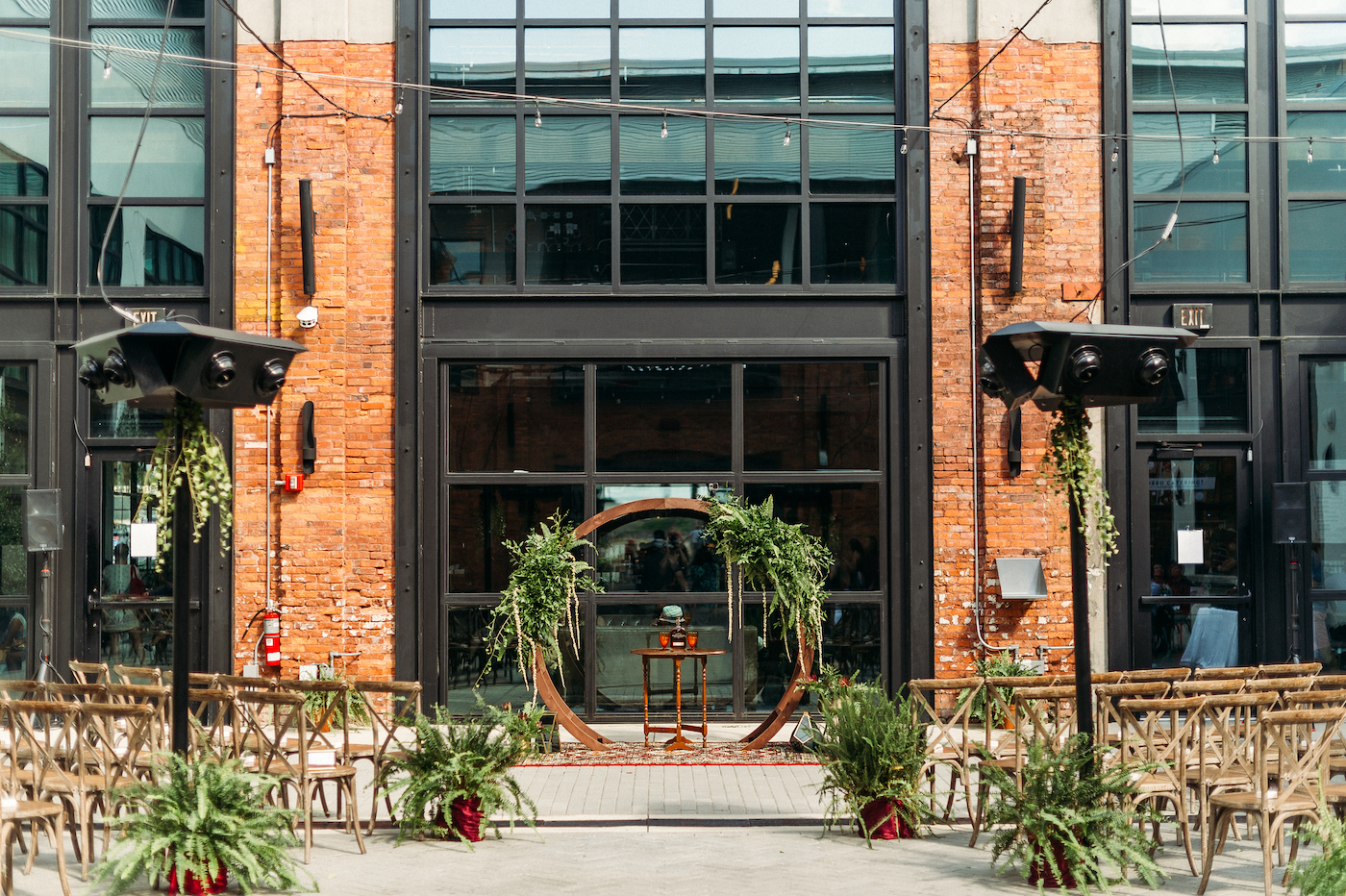 Tampa Wedding Armature Works Courtyard Ceremony with Canopy String Lights and Wood Cross Back Chairs and Round Moon Arch | Ceremony Aisle with Potted Ferns and Greenery Wedding Floral Arrangements | Outdoor Tampa Weddings