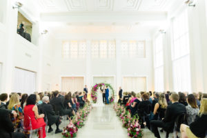 Indoor Downtown Tampa Wedding Ceremony at Tampa Wedding Venue The Vault | Bright Colorful Florida Wedding Ceremony Floral Circle Moon Round Arch with Pink Roses and Purple Orchids and Greenery | Ceremony Aisle Flowers with Blush Pink Gladiolas | Clear Ghost Chairs from Kate Ryan Event Rentals