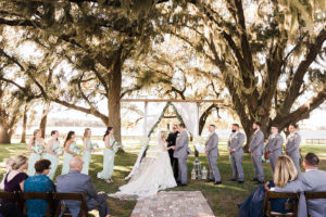 Rustic Elegant Outdoor Wedding Ceremony and Decor, Florida Bride and Groom Exchange Vows Under Wooden Arch, Crucifix Cross Backdropm White Draping with Eucalyptus Leaf Greenery, Vintage Ivory Bird Cages, Bridesmaids in Long Sage Green Dresses | Tampa Bay Luxury Wedding Planner and Floral Designer John Campbell Weddings | Covington Farm
