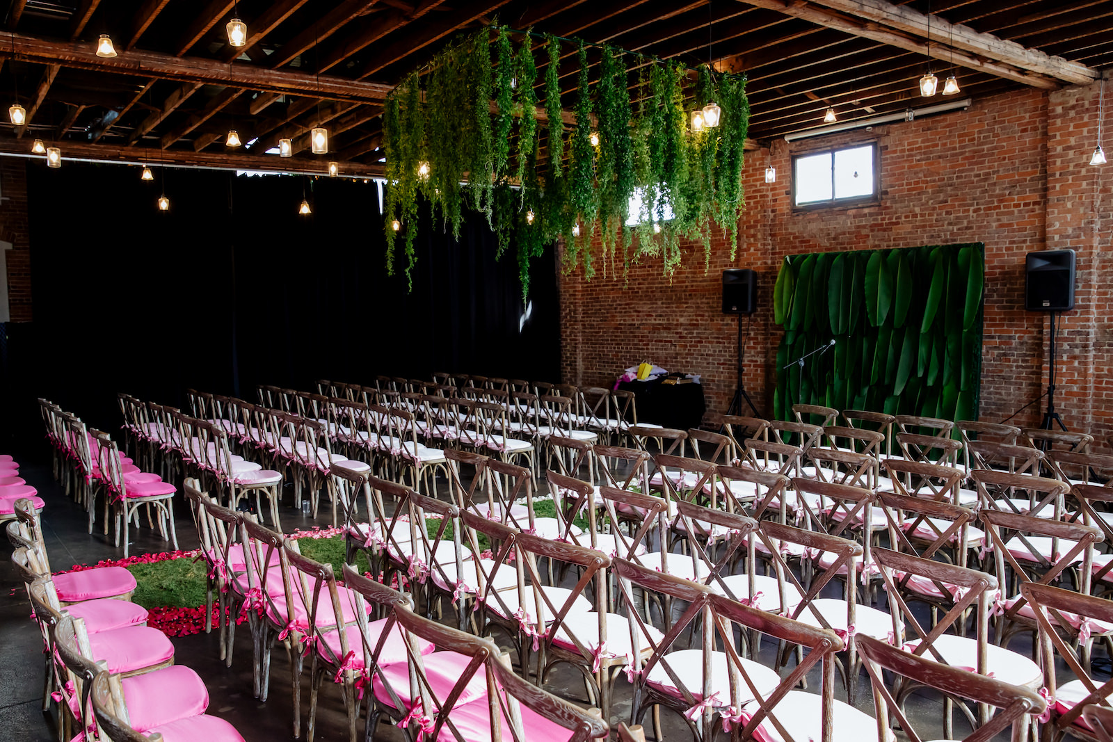 Modern Wedding Ceremony Backdrop with Tropical Palm Leaves and Moss Aisle Runner with Ombre Pink and White Rose Petals | Wood Cross Back Chairs and Historic Brick Walls | Suspended Greenery Garland Ceiling Installment Wedding Floral Chandelier