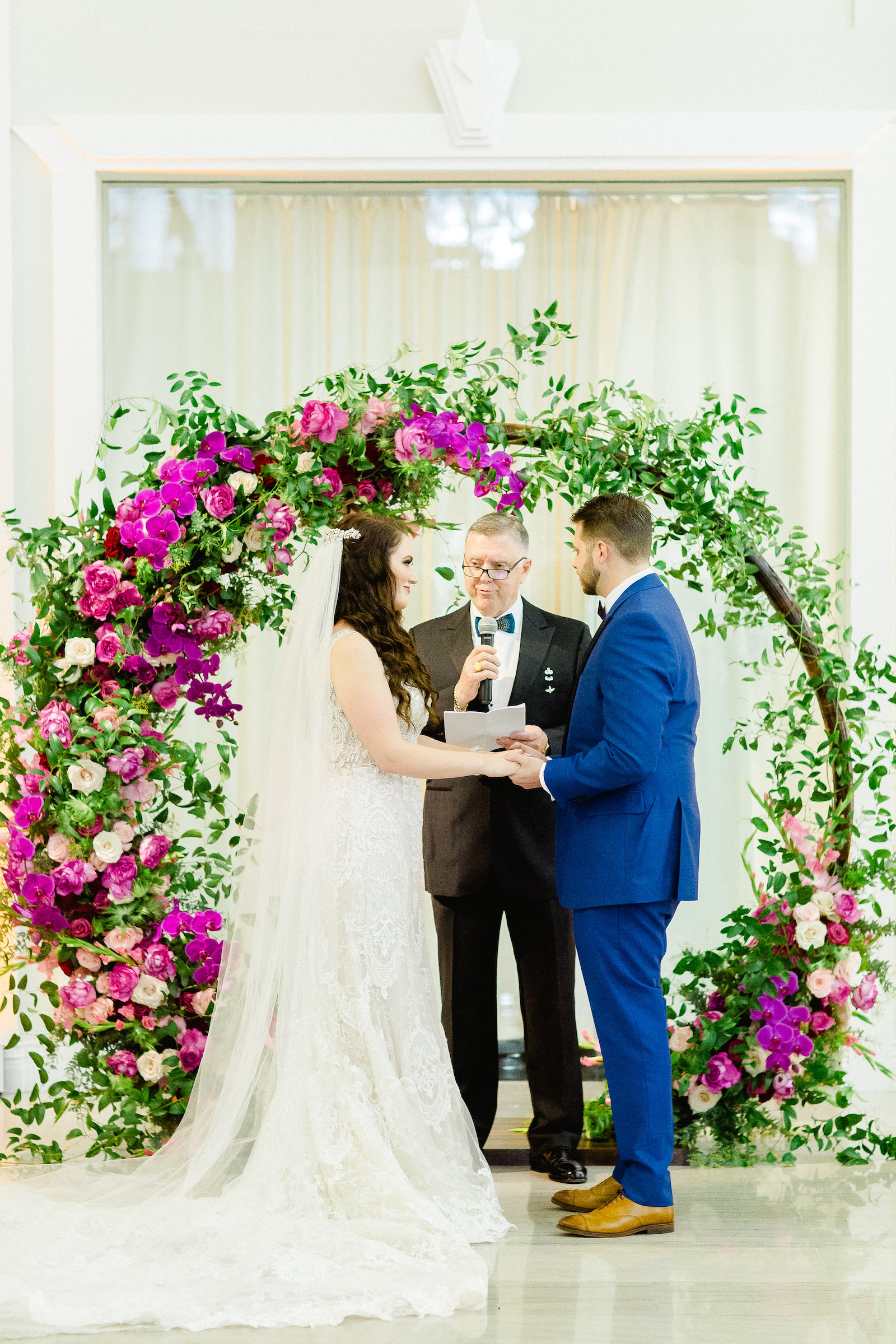 Bride and Groom Exchanging Vows at Indoor Downtown Tampa Wedding Ceremony at Tampa Wedding Venue The Vault | Bright Colorful Florida Wedding Ceremony Floral Circle Moon Round Arch with Pink Roses and Purple Orchids and Greenery | Groom Wearing Classic Blue Suit