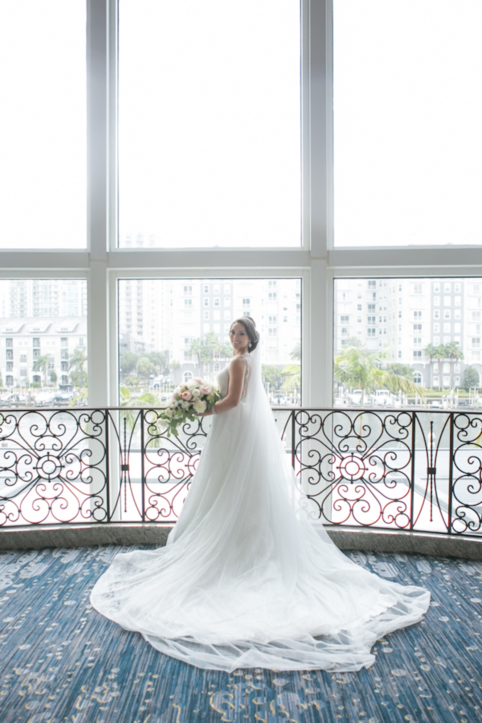 Indoor Downtown Tampa Hotel Window Bridal Portrait at Marriott Water Street | V Neck Illusion Lace A Line Ballgown Wedding Dress with Rhinestone Waist Band and Cathedral Veil | Blush Pink and White Roses and Peonies Wedding Bridal Bouquet