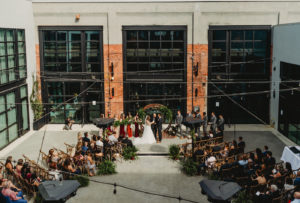 Tampa Wedding Armature Works Courtyard Ceremony with Canopy String Lights and Wood Cross Back Chairs and Round Moon Arch | Ceremony Aisle with Potted Ferns and Greenery Wedding Floral Arrangements