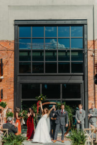 Bride and Groom Pronounced Husband and Wife After Ceremony | Tampa Wedding Armature Works Courtyard Ceremony with Canopy String Lights and Wood Cross Back Chairs and Round Moon Arch | Ceremony Aisle with Potted Ferns and Greenery Wedding Floral Arrangements | Dewitt for Love Photography