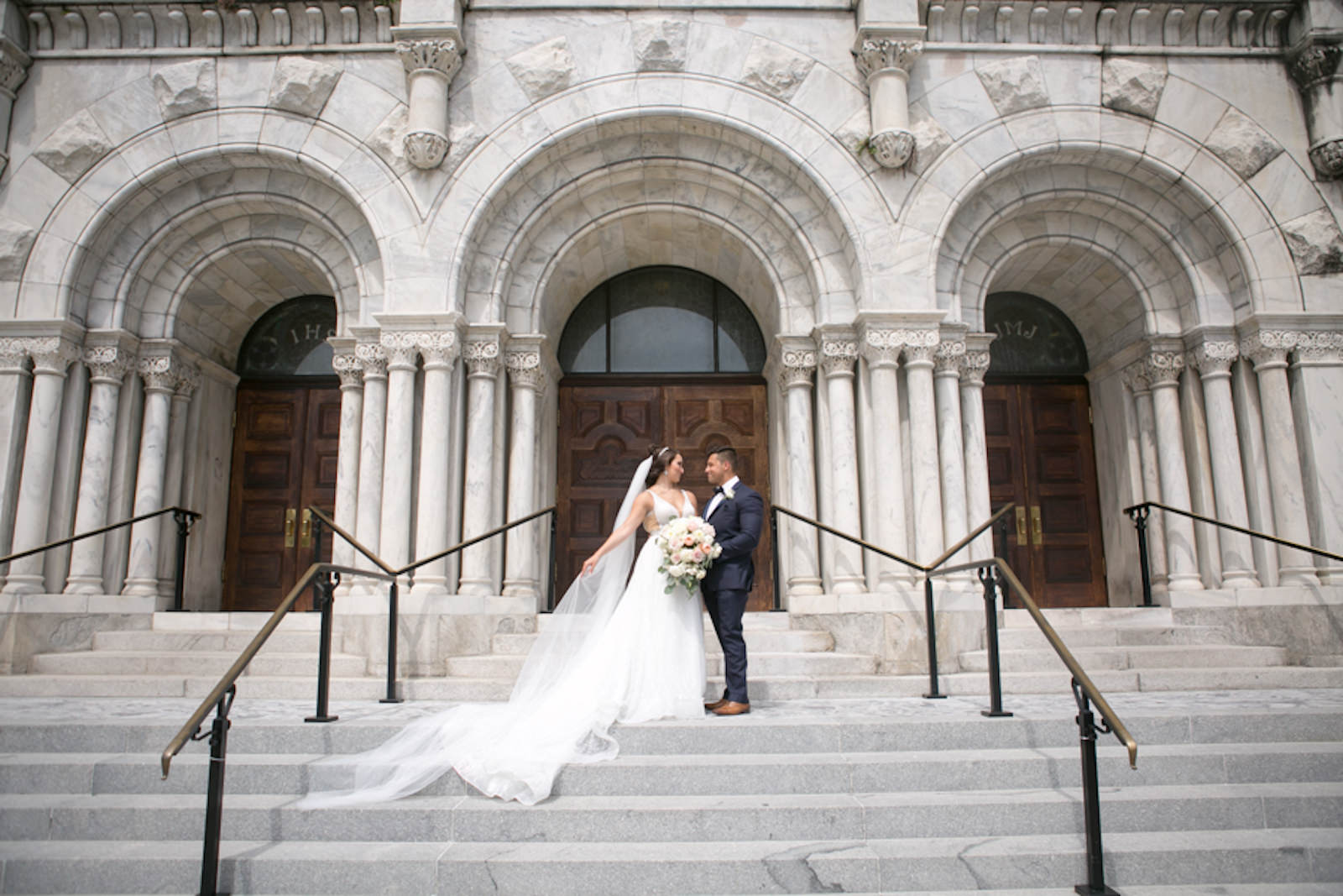 Outdoor Downtown Tampa Bride and Groom Portrait at Traditional Catholic Ceremony Cathedral Church Staircase | V Neck Illusion Lace A Line Ballgown Wedding Dress with Rhinestone Waist Band and Cathedral Veil | Groom in Classic Navy Blue Suit with Bow Tie | Blush Pink and Ivory Roses and Peonies Bridal Bouquet with Eucalyptus Greenery | Carrie Wildes Photography