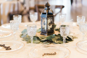 Rustic Elegant Candle Centerpieces, Antique Lantern on Wooden Stand, Surrounded by White Roses In Eucalyptus Leaf Greenery, Crystal Stemware and Custom Laser Cut Wood Place Cards | Florida Wedding Planner and Floral Designer John Campbell Weddings | Florida Wedding Caterer Olympia Catering | Table Linens Kate Ryan Event Rentals