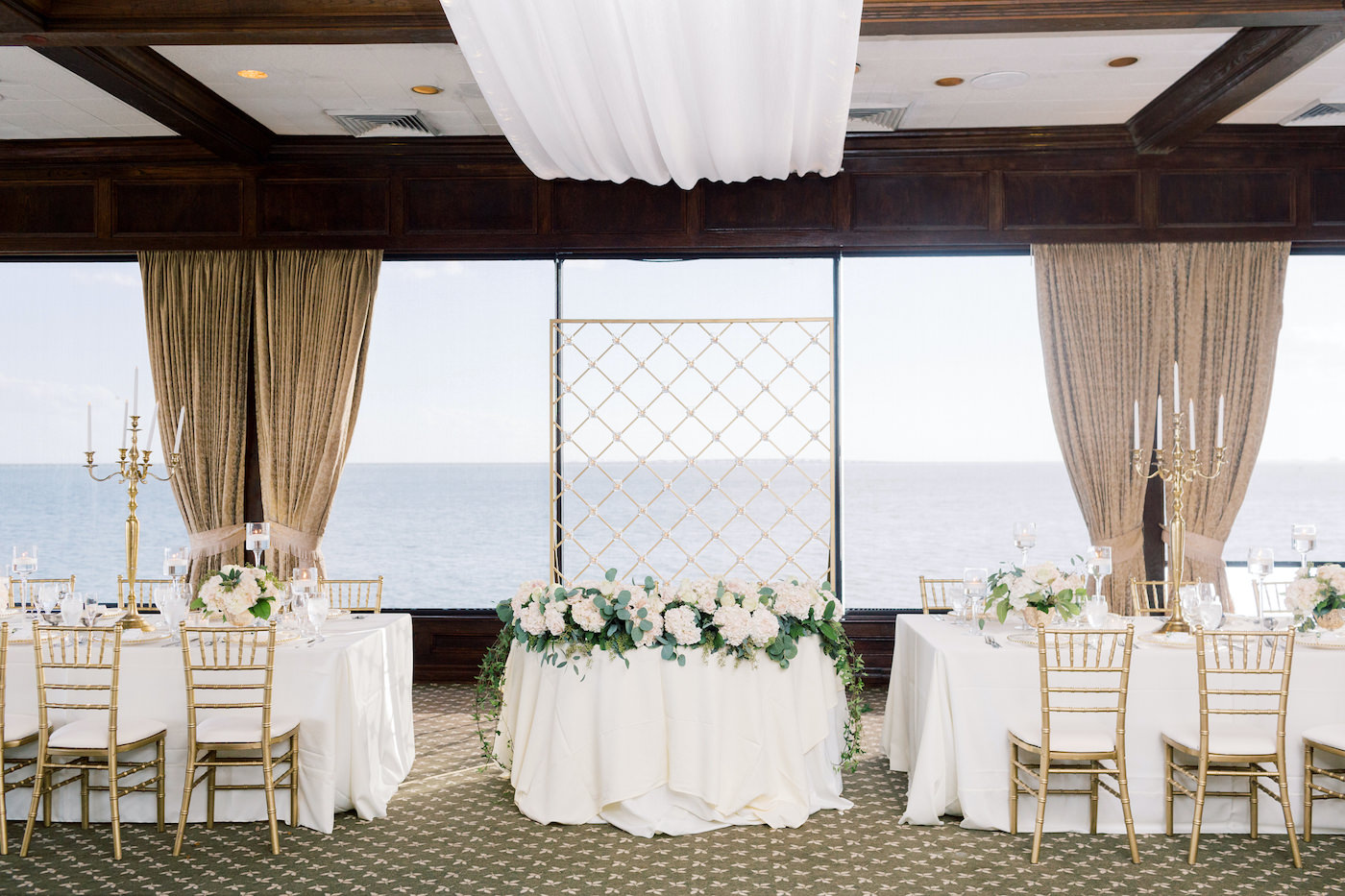 Classic Romantic Wedding Reception Decor, Sweetheart Table with Criss Cross Gold Wall Backdrop and Greenery and White Floral Garland, Gold Chiavari Chairs, Tall Candelabras | Tampa Bay Wedding Planner Special Moments Event Planning | Wedding Florist and Candelabra Rental Gabro Event Services | Waterfront Wedding Reception Venue The Rusty Pelican