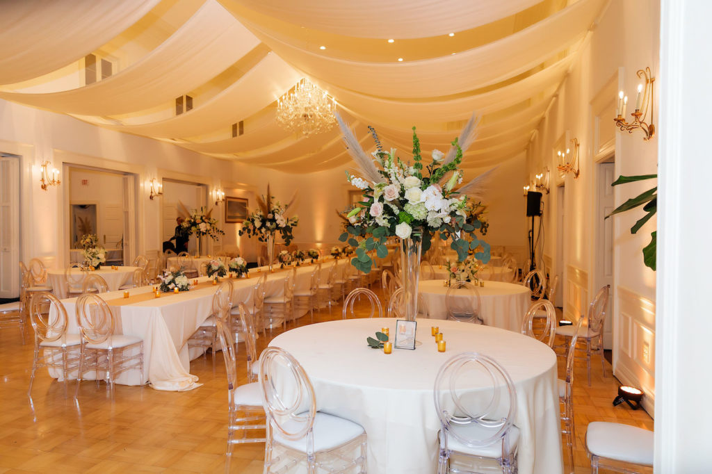 Whimsical Romantic Wedding Reception Decor, White Draped Ceiling, Crystal Chandelier, Round Tables with White Linens and Long Feasting Table, Acrylist Ghost Chairs with White Cushions, Tall Glass Vases with Lush Blush Pink and White Roses, Eucalyptus Greenery, Hydrangeas, Pampas Grass Floral Tall Centerpiece | Tampa Wedding Planner Breezin' Weddings | Wedding Venue Tampa Yacht and Country Club