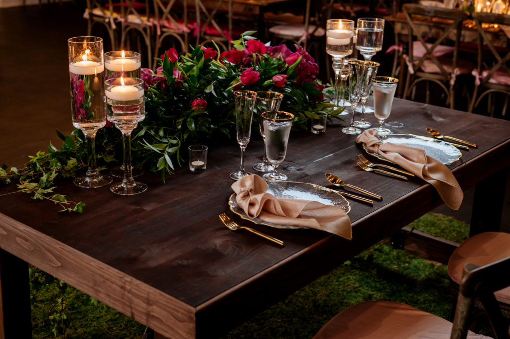 Wood Farm Wedding Reception Sweetheart Table with Greenery and Pink Roses Arrangement and Floating Candles | Wedding Reception Place Setting with Gold Rimmed Glassware and Gold Flatware with Gold Rim Glass Charger Plate by Kate Ryan Event Rentals | Kate Ryan Event Rentals