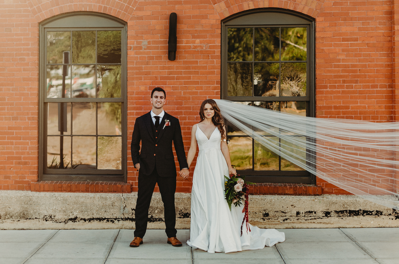 Bride and Groom Outdoor Wedding Portrait Veil Shot In Front Of Brick Wall | Charcoal Grey Wool Groom Suit with Burgundy Maroon Red Neck Tie | Wild Boho Loose Organic Greenery Bridal Bouquet with Ferns Blush Pink Roses and Protea and Red Amaranthus | Dewitt for Love Photography