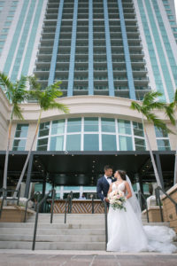 Outdoor Downtown Tampa Bride and Groom Portrait at Marriott Water Street Hotel | V Neck Illusion Lace A Line Ballgown Wedding Dress with Rhinestone Waist Band and Cathedral Veil | Groom in Classic Navy Blue Suit with Bow Tie | Blush Pink and Ivory Roses and Peonies Bridal Bouquet with Eucalyptus Greenery