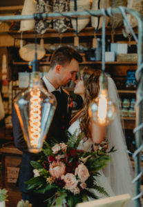 Bride and Groom Indoor Wedding Portrait with Edison Bulbs | Charcoal Grey Wool Groom Suit with Burgundy Maroon Red Neck Tie | Wild Boho Loose Organic Greenery Bridal Bouquet with Ferns Blush Pink Roses and Protea and Red Amaranthus | Dewitt for Love Photography