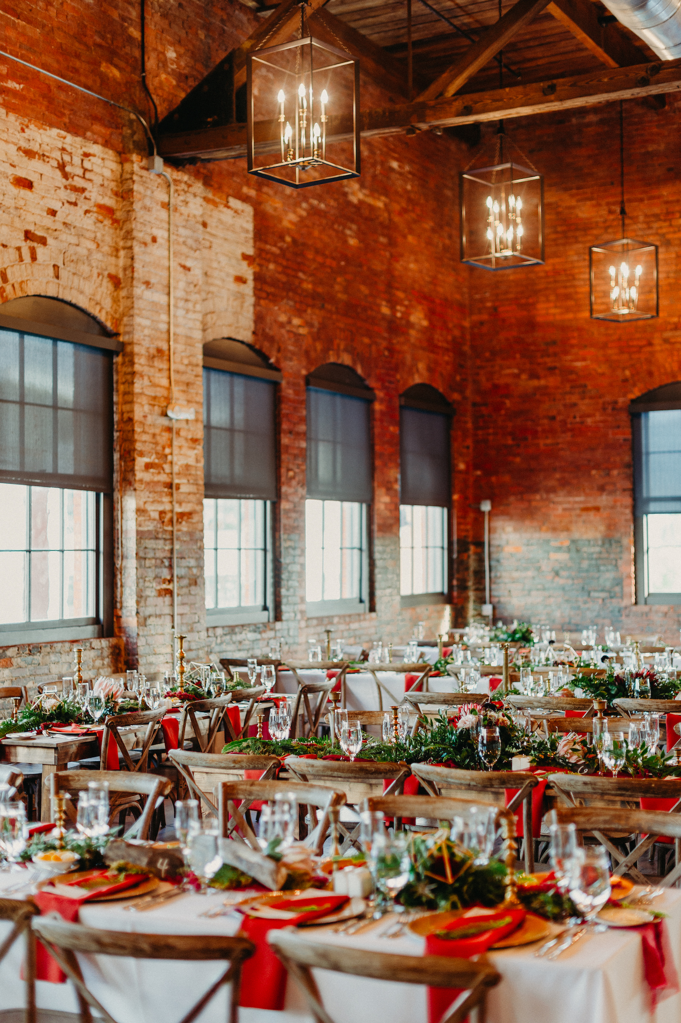 Tampa Wedding Reception at Historic Building Armature Works with Brick Walls and Industrial Chic Chandeliers | Long Feasting Tables with Wood Cross Back Chairs and Greenery Garland Runners with Burgundy Maroon Red Napkins