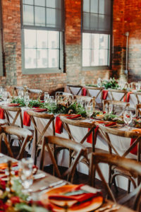 Tampa Wedding Reception at Historic Building Armature Works with Brick Walls and Industrial Chic Chandeliers | Long Feasting Tables with Wood Cross Back Chairs and Greenery Garland Runners with Burgundy Maroon Red Napkins