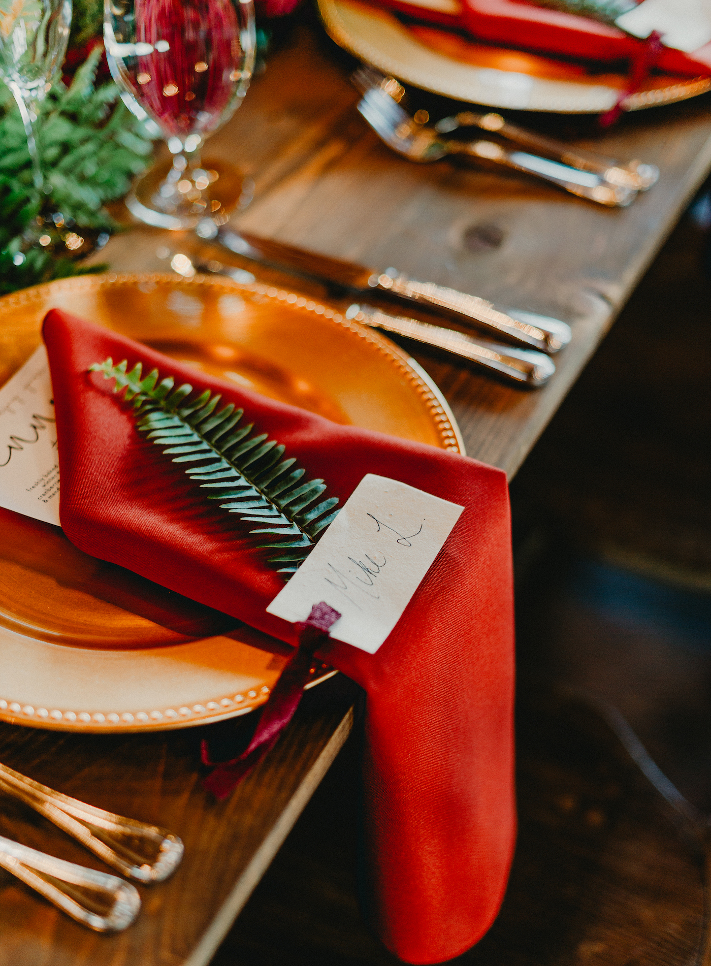 Wedding Reception Wood Feasting Farm Tables | Wedding Place Settings with Gold Charger Plates and Gold Flatware with Burgundy Maroon Red Napkin and Fern Greenery with Menu Card and Place Card