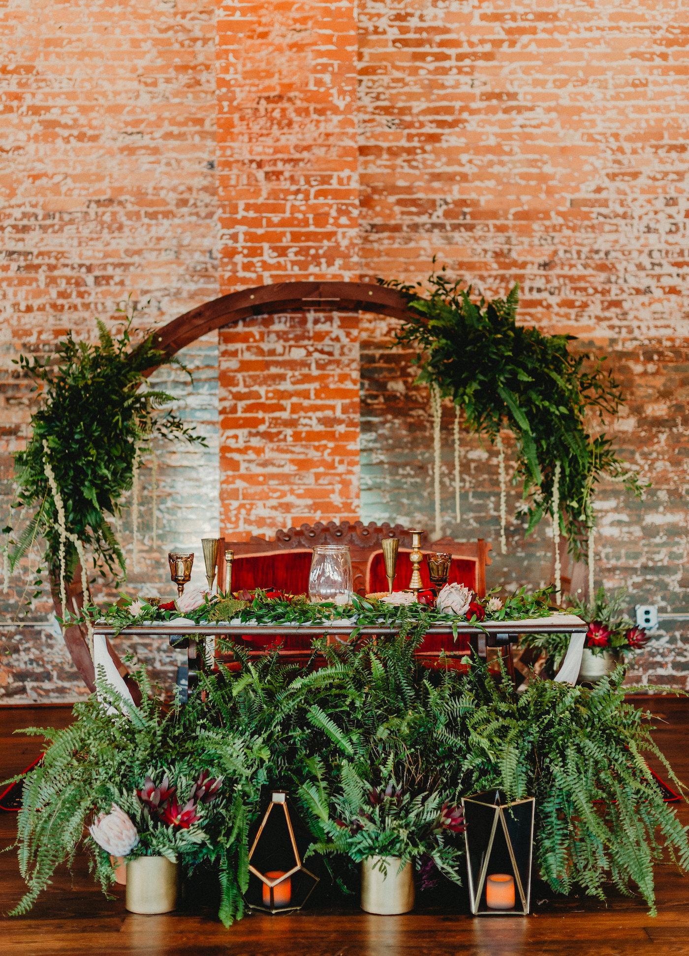 Bride and Groom Wedding Sweetheart Table with Red Vintage Loveseat | Round Moon Arch with Greenery Wedding Floral Arrangements with Potted Ferns and Gold Geometric Candle Lanterns