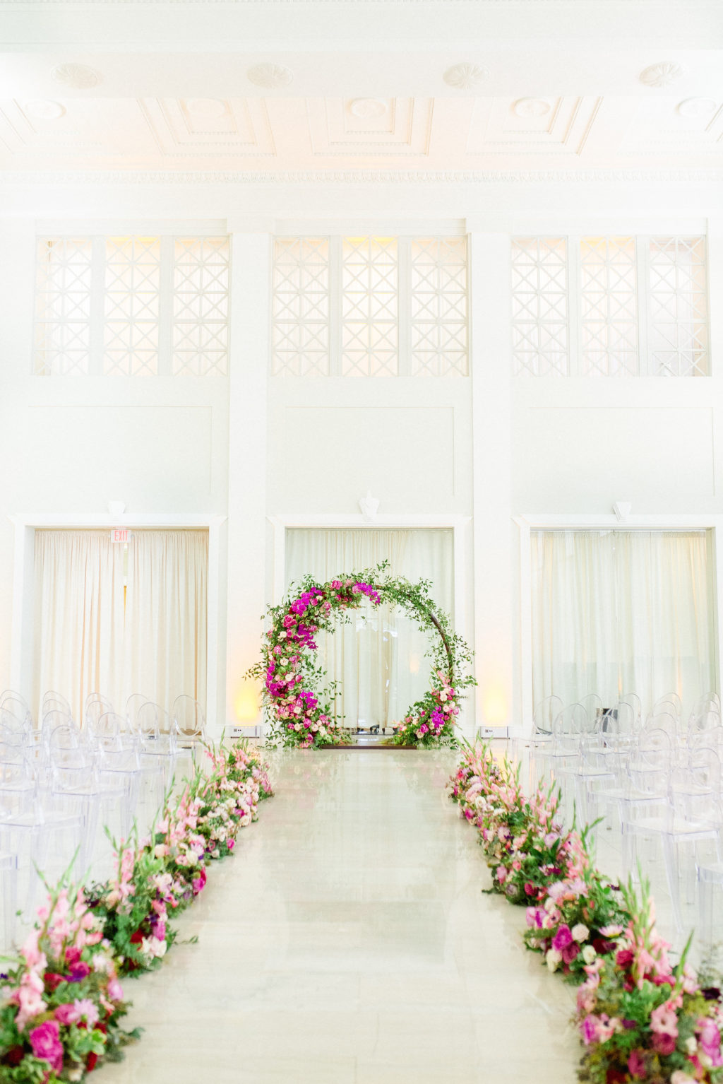Indoor Downtown Tampa Wedding Ceremony at Tampa Wedding Venue The Vault | Bright Colorful Florida Wedding Ceremony Floral Circle Moon Round Arch with Pink Roses and Purple Orchids and Greenery | Ceremony Aisle Flowers with Blush Pink Gladiolas | Clear Ghost Chairs from Kate Ryan Event Rentals
