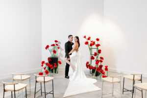 Modern Clean Indoor Ceremony Space at Tampa Wedding Venue Hyde House | Black White and Red Ceremony Backdrop with Red Amaryllis and Black Columns | Dramatic Modern Ines di Santo High Slit Wedding Dress by Isabel Oneil Bridal | Groom in All Black Suit | Black and White Birch Ceremony Chairs