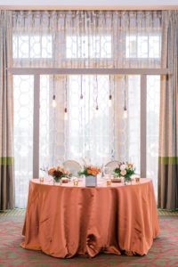 Copper Orange Wedding Sweetheart Table Linens with Gold Votive Candles and Suspended Edison Bulb Lights Arch | Fall Autumn Wedding Centerpiece with Orange and Peach Roses and Eucalyptus Greenery