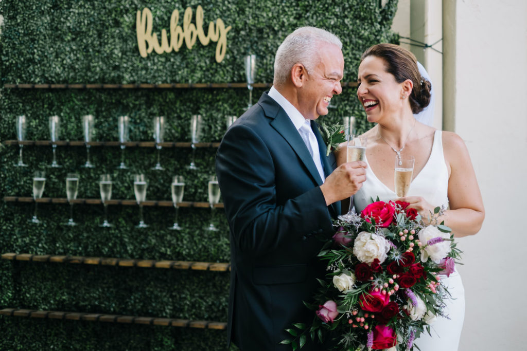Tampa Bay Bride and Groom Cheers in front of Champagne Wall, Greenery Boxwood Bubbly Wall Rental, Bride Holding Vibrant Jewel Toned Floral Bouquet with Bright Pink Roses, Ivory, Purple, and Blue Flowers | Florida Wedding Planner Breezin' Weddings | South Tampa Outside The Box Event Rentals