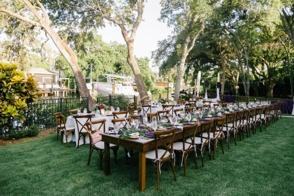 Romantic Outdoor Backyard Reception on the water, Long Wooden Feasting Tables with Farmhouse Chairs, Jeweled Tone Tablescape with Greenery Garland, Vibrant Pink Roses, Purple Linens, Gold Chargers | South Tampa Wedding Planner Breezin' Weddings | Beach Park Wedding Rentals Outside the Box Event Rentals | Tampa Bay Wedding DJ Breezin' Entertainment