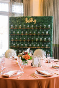 Wedding Champagne Wall Greenery Boxwood Hedge with Glasses of Bubbly by Outside the Box Event Rentals | Copper Orange Wedding Reception Table Linens with Gold Beaded Glass Charger Plates and Gold Flatware | Fall Autumn Wedding Centerpiece with Orange and Peach Roses and Pampas Grass
