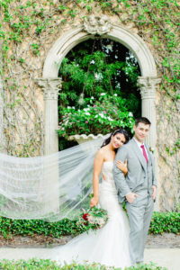 Outdoor Bride and Groom Portrait Veil Shot in Fairy Tale Garden Ivy Walls | Mermaid Fitted Lace Sweetheart Wedding Gown with Tulle Skirt and Sheer Illusion Panels with Long Cathedral Veil from Tampa Dress Shop Isabel Oneil Bridal Collection | Deep Red Burgundy and Blush Pink Rose Bouquet | Groom Wearing Classic Silver Grey Suit with Burgundy Neck Tie