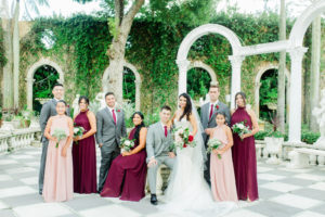 Outdoor Wedding Party Group Portrait Fairy Tale Garden | Mermaid Fitted Lace Sweetheart Wedding Gown with Tulle Skirt and Sheer Illusion Panels with Long Cathedral Veil from Tampa Dress Shop Isabel Oneil Bridal Collection | Deep Red Burgundy and Blush Pink Rose Bouquet | Groom and Groomsmen Wearing Classic Silver Grey Suit with Burgundy Neck Tie | Blush Pink and Burgundy Long Chiffon Bridesmaid Dresses from Tampa Dress Shop Nikki's Glitz & Glamour Boutique