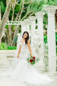 Outdoor Bridal Portrait in Fairy Tale Sculpture Garden with Columns | Mermaid Fitted Lace Sweetheart Wedding Gown with Tulle Skirt and Sheer Illusion Panels with Long Cathedral Veil from Tampa Dress Shop Isabel Oneil Bridal Collection | Deep Red Burgundy and Blush Pink Rose Bouquet
