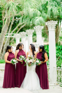Outdoor Bridal Party Portrait in Fairy Tale Sculpture Garden with Columns | Mermaid Fitted Lace Sweetheart Wedding Gown with Tulle Skirt and Sheer Illusion Panels with Long Cathedral Veil from Tampa Dress Shop Isabel Oneil Bridal Collection | Deep Red Burgundy and Blush Pink Rose Bouquet | Long Burgundy Deep Red Chiffon Bridesmaid Dresses by JJ's House from Tampa Dress Shop Nikki's Glitz & Glam Boutique