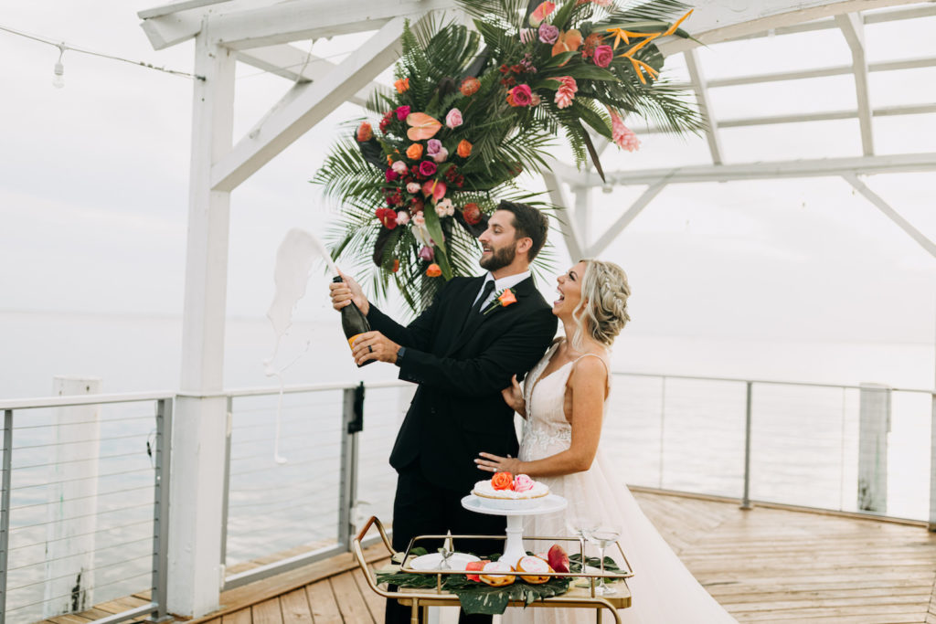 Florida Bride in Boho Flowy Dress with Groom Popping Champagne Bottle on Waterfront Pier, Tropical Colorful Floral Arrangement with Palm Tree Leaves, Pink, Purple, Orange Roses | Tampa Bay Wedding Venue The Godfrey | Wedding Florist Brides N Blooms | Wedding Planner Elope Tampa Bay | Wedding Dress NIkki's Glitz and Glam | Wedding Hair and Makeup Femme Akoi Beauty Studio | Amber McWhorter Photography