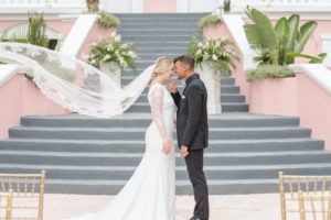 Romantic Bride in High Scoop Neckline and Lace Long Sleeve Wedding Dress and Lace Trim Veil Blowing in Wind, Groom in All Black Suit Intimate Photo | Don Cesar Styled Wedding | Tampa Bay Wedding Dress Shop Truly Forever Bridal