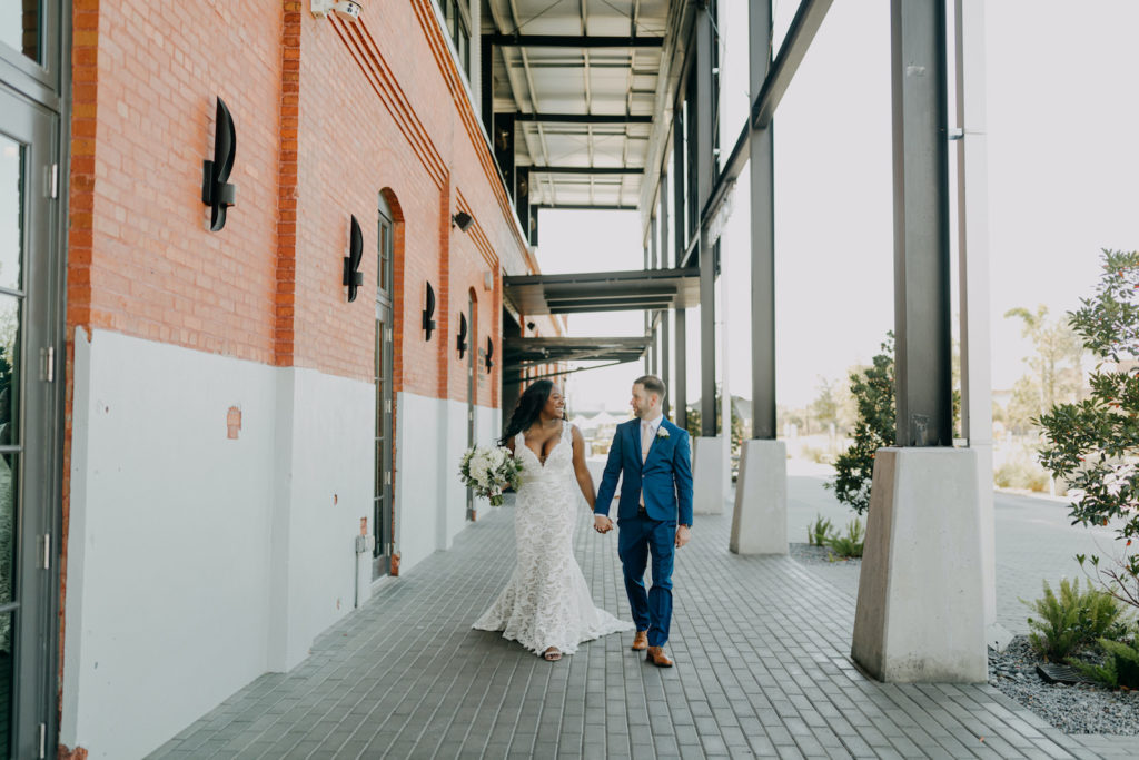 Bride and Groom Outdoor Downtown Tampa Heights Riverwalk Wedding Portrait at Armature Works | Ivory Lace over Champagne Lining Bridal Gown V Neck Sheer Illusion Plunge Wedding Dress by BHLDN Wtoo Watters | Groom Wearing Modern Blue Suit | Classic Round Ivory Rose and Hydrangea Bridal Bouquet with Eucalyptus Greenery | Amber McWhorter Photography