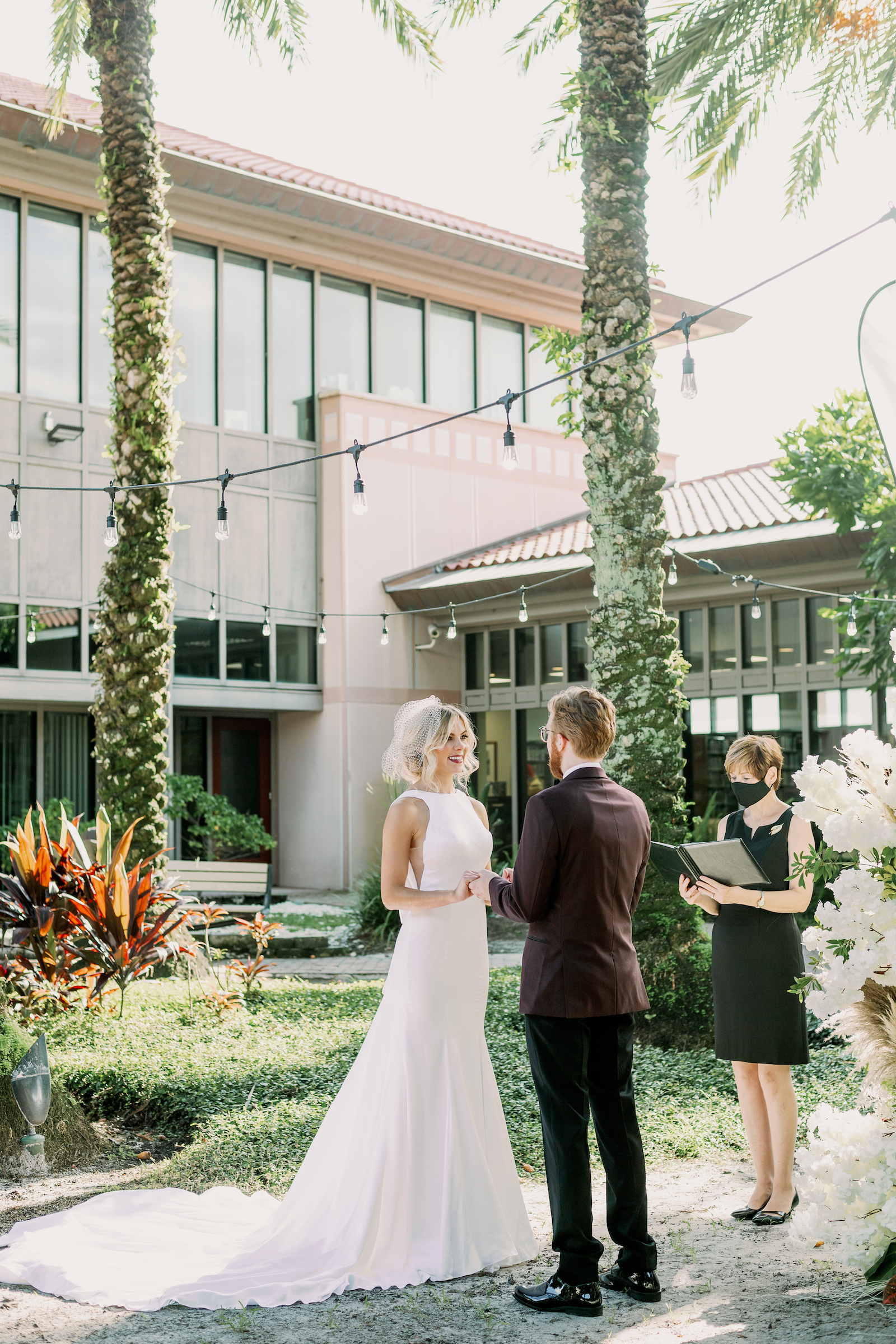 Bride and Groom Outdoor Atrium Intimate COVID Elopement Micro Wedding Ceremony at St. Pete Wedding Venue The Poynter Institute | LoAdoro Bridal Sleek Fitted Modern Wedding Dress Bridal Gown with Birdcage Veil from Tampa Bridal Shop Truly Forever Bridal | Groom in Modern Suit with Dark Red Maroon Jacket with Black Satin Lapel and Bow Tie | Tampa Wedding Officiant A Wedding With Grace