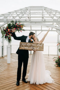 Florida Bride in Boho Flowy Dress with Groom Kissing Holding We Do We Did We Eloped Sign, on Waterfront Pier, Tropical Colorful Floral Arrangement with Palm Tree Leaves, Pink, Purple, Orange Roses | Tampa Bay Wedding Venue The Godfrey | Wedding Florist Brides N Blooms | Wedding Planner Elope Tampa Bay | Wedding Dress NIkki's Glitz and Glam | Wedding Hair and Makeup Femme Akoi Beauty Studio | Amber McWhorter Photography