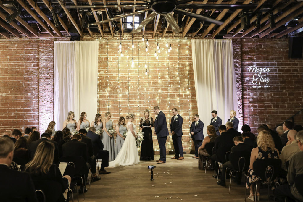 Modern, Simplistic Indoor Tampa Bay Wedding Ceremony, Downtown St. Petersburg Bride and Groom Exchange Vows With Exposed Brick Walls and Romantic String Lighting in Backdrop, White Draping, and Light Gobo of Custom Monogram | Florida Historic Wedding Venue NOVA 535 | St. Petersburg Wedding Photographer Lifelong Photography Studios