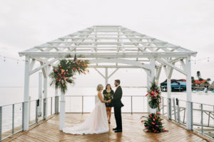 Groom and Bride Exchanging Wedding Vows Wearing Romantic Flowy Boho Wedding Dress, Open V Back on Waterfront Pier, Tropical Colorful Floral Arrangements, Styled Shoot | Tampa Bay Wedding Venue The Godfrey | Wedding Planner Elope Tampa Bay | Wedding Dress Nikki's Glitz and Glam | Wedding Florist Brides N Blooms | Amber McWhorter Photography