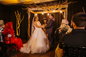 Halloween Bride and Groom Exchanging Vows During Ceremony Under Wooden Branches Arch and Black Linen Draping | Tampa Bay Wedding Planner UNIQUE Weddings and Events | Wedding Rentals A Chair Affair and Wedding Linens Over the Top Rental Linens | Wedding Dress Truly Forever Bridal