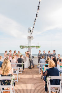 Tampa COVID Wedding at Yacht Starship Venue | Bride and Groom First Kiss during Waterfront Wedding Ceremony Ship Boat Deck with White Folding Garden Chairs and Greenery Garland | Blush Pink and White and Grey Silver Wedding | Blush Pink Bridesmaid Dresses by Lulu's | Groom and Groomsmen in Silver Grey Suit Tux