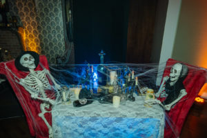 Creepy Halloween Wedding Reception Decor, Two Skeletons Covered in Cobwebs Sitting in Red Velvet Chairs at Table with Lace Linen | Tampa Bay Wedding Planner UNIQUE Weddings and Events | Wedding Chair Rentals A Chair Affair | Wedding Linen Rentals Over the Top Rental Linens