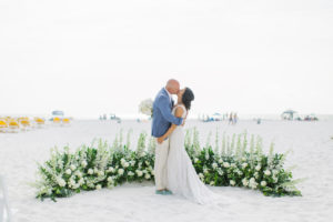 Classic bride and groom on sandy beach with half circle floral arrangement of tall white flowers, roses and greenery, groom wearing pale blue suit jacket and cream linen pants | Tampa wedding planner Parties A'la Carte